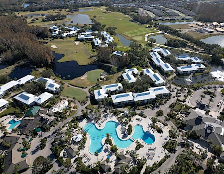 The image shows an aerial view of a resort with pools, green spaces, and multiple residential buildings.