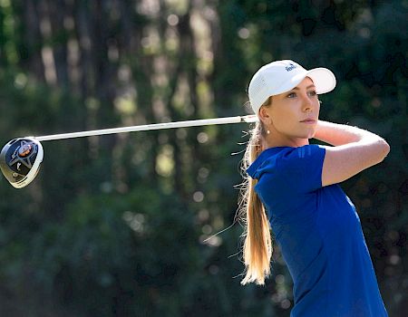 A person is playing golf, wearing a blue shirt and white cap, mid-swing with a golf club against a backdrop of trees, focused on the shot.